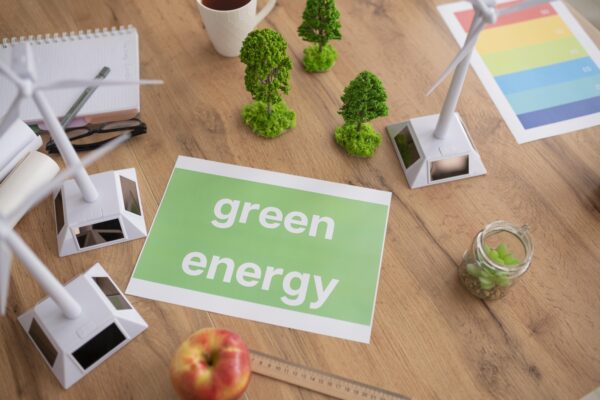 grünes Blatt Papier auf einem Holztisch mit der weißen Schrift "Green energy". Auf dem Tisch sind kleine grüne Modellbäume und Modellwindräder zu sehen.